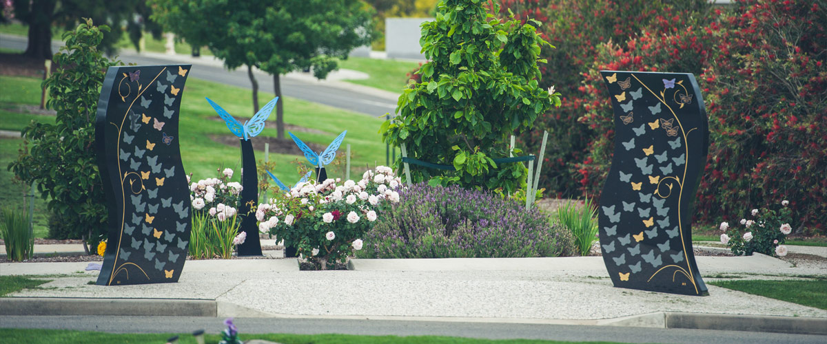 Three blue metal butterflies in a colourful garden, with memorial wall in cemetery